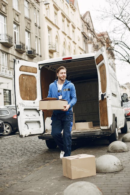 A man wearing a blue jacket and dark trousers is engaged in the loading process during a house move in Islington. He is holding a medium-sized cardboard box with packing tape, preparing to load it into the rear of a white van with open doors, which is parked on a cobblestone street. The van’s interior is visible, showing additional cardboard boxes and packing materials stacked inside, with some boxes still on the ground nearby. The scene takes place in an urban residential area with multi-storey, cream-coloured buildings featuring window air conditioning units and balconies. Several other parked cars are visible along the street. The environment appears to be late autumn or winter, as indicated by the bare tree branches. This captured moment demonstrates a professional furniture transport and packing process, typical of house removals services offered by Islington Man and Van, focusing on efficient loading and home relocation logistics.