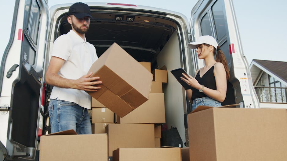 A man and a woman are standing at the open rear of a white moving van, participating in a home relocation process. The man, dressed in a white polo shirt, jeans, and a black cap, is holding a medium-sized cardboard box, likely preparing to load or unload it from the vehicle, which is filled with several other cardboard boxes of various sizes. The woman, wearing a sleeveless black top, high-waisted jeans, and a white cap, is looking at a tablet, possibly reviewing a moving checklist or inventory. The inside of the van reveals a partly visible interior with more boxes, and it is parked on a residential street with a house visible in the background. Items are being packed or staged for transport, with some boxes placed on the pavement and gentle natural lighting illuminating the scene. This image relates to house removals, furniture transport, and packing and moving services offered by Islington Man and Van at Angel Station, N1.