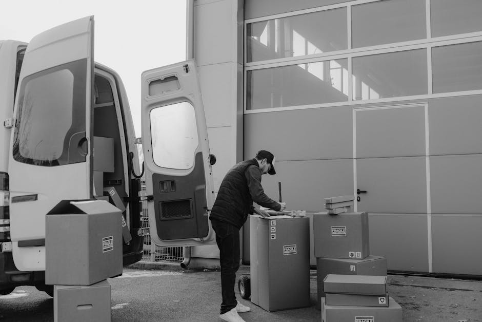 A man wearing a blue jacket and dark trousers is engaged in the loading process during a house move in Islington. He is holding a medium-sized cardboard box with packing tape, preparing to load it into the rear of a white van with open doors, which is parked on a cobblestone street. The van’s interior is visible, showing additional cardboard boxes and packing materials stacked inside, with some boxes still on the ground nearby. The scene takes place in an urban residential area with multi-storey, cream-coloured buildings featuring window air conditioning units and balconies. Several other parked cars are visible along the street. The environment appears to be late autumn or winter, as indicated by the bare tree branches. This captured moment demonstrates a professional furniture transport and packing process, typical of house removals services offered by Islington Man and Van, focusing on efficient loading and home relocation logistics.
