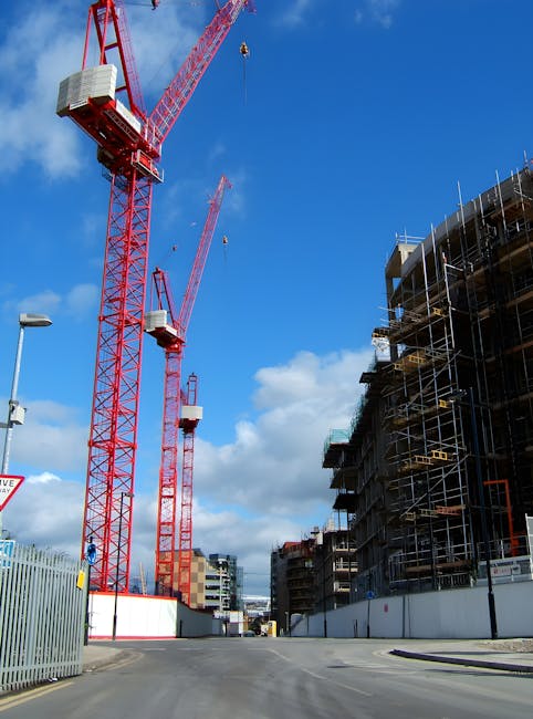 A construction site on a clear day showing two large red tower cranes with long booms extending high into the blue sky, equipped with counterweights and operator cabins. The site features a multi-storey building under construction, surrounded by scaffolding with metal poles and wooden planks, and partially covered by protective netting. The area is enclosed by metal fencing, and the scene is illuminated by natural daylight, indicating active construction and lifting operations typical in building projects. The image provides context for logistics and equipment used in construction, paralleling home relocation and furniture transport activities handled by companies like Islington Man and Van, especially during moves involving building sites or new property developments.