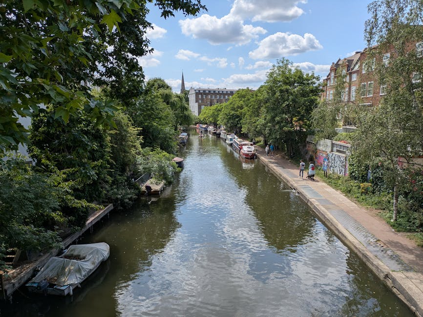 A narrow canal in an urban setting during daytime, lined with dense green trees on both sides. On the right, there is a paved walkway with a few pedestrians walking along it, and parked cars are visible further down the street. On the left, boats are moored along the water's edge, with one boat covered in a grey tarp. The water reflects the blue sky and scattered clouds overhead. In the background, brick residential buildings and a church steeple are visible, with lush foliage and a mix of modern and historic architecture. This scene reflects a typical house relocation neighborhood near Islington Town Hall, where furniture transport and moving logistics may take place under the supervision of companies like Islington Man and Van.