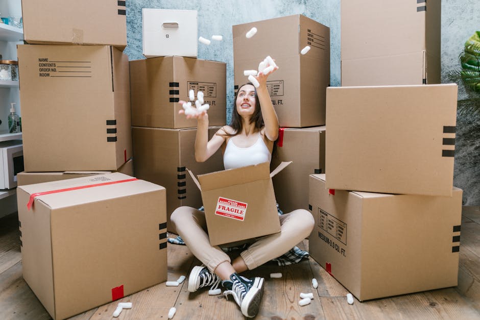 A young woman sitting cross-legged on a wooden floor surrounded by large cardboard moving boxes, some sealed with red packing tape and others open, with packing materials like white packing peanuts scattered around her. She is inside a room with a textured blue wall, a tall green plant in the background, and a small shelving unit with containers on the left. The woman is holding and tossing packing peanuts into the air, smiling, and wearing casual clothing including a white tank top, beige pants, and black and white sneakers. The scene captures the packing stage of a home relocation, with a focus on the process of organizing and preparing belongings for moving, supported by the presence of sturdy cardboard boxes and packing materials. This visual demonstrates typical packing activities during furniture transport and home removals, highlighting the importance of careful packing and logistics, as performed by Islington Man and Van.