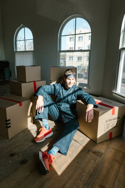 A person dressed in casual clothing, including a blue jacket, dark pants, and bright orange sneakers, is sitting on the wooden floor inside a room with large, arched windows. The individual is surrounded by several packed cardboard boxes, some sealed with red tape, suggesting they are ready for a home relocation. The boxes vary in size and are stacked or placed nearby, indicating a packing or moving process in progress. Natural light from the windows illuminates the space, highlighting the person's relaxed pose and the interior environment, which features light-colored walls and a clean, uncluttered floor. This scene exemplifies the packing and moving stage of house removals, as well as the logistical efforts involved, often managed by professional services like Islington Man and Van.