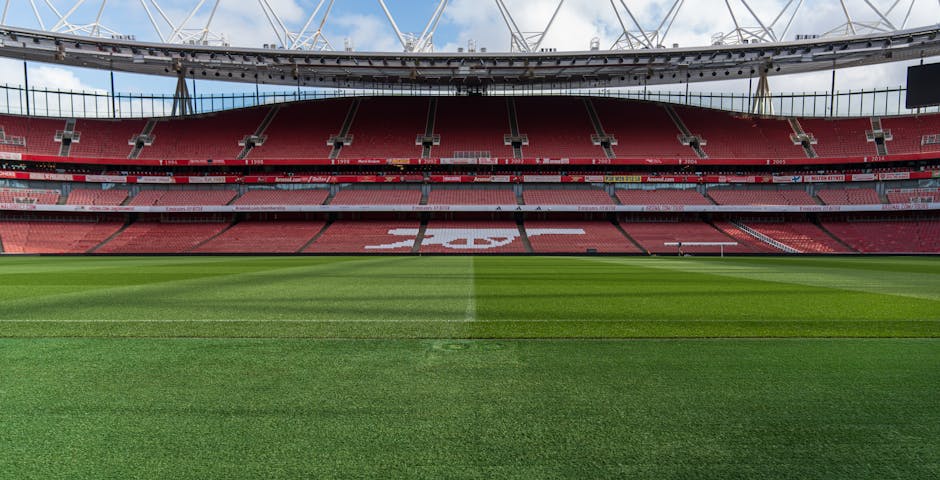 The image shows the interior of a large, modern football stadium with a well-maintained grass pitch marked for a match, viewed from the stands and looking towards the seating area. The stadium features multiple tiers of red seating, with the upper tier displaying the sponsor signage. Overhead, a semi-transparent roof structure supported by white steel beams covers the seating sections. The lighting appears to be natural daylight, with some cloud cover visible in the sky above. The focus is on the playing field and the seating environment, typical of venues used for football matches, events, and sports gatherings. There are no household or moving-related objects present in the image, and no furniture, boxes, or moving equipment are visible, indicating this is a sports stadium setting rather than a home or relocation site.