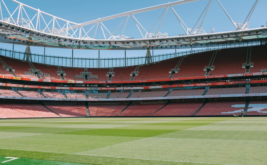 The image shows an empty football stadium during daylight hours, with a well-maintained green pitch in the foreground and tiered rows of red and pink seating areas behind. Above the seating area, there are large advertising banners displaying various sponsor names. The stadium's roof structure, consisting of white steel beams and trusses, extends over the seating sections, supporting a partial roof cover. The sky is clear with no clouds, and the lighting suggests bright, natural sunlight. This setting is unrelated to house removals or moving services, but the detailed structural elements and open space reflect the scale and design of a professional sports arena.