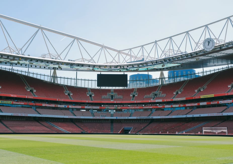 The image shows the interior of a large, modern football stadium with a well-maintained grass pitch marked for a match, viewed from the stands and looking towards the seating area. The stadium features multiple tiers of red seating, with the upper tier displaying the sponsor signage. Overhead, a semi-transparent roof structure supported by white steel beams covers the seating sections. The lighting appears to be natural daylight, with some cloud cover visible in the sky above. The focus is on the playing field and the seating environment, typical of venues used for football matches, events, and sports gatherings. There are no household or moving-related objects present in the image, and no furniture, boxes, or moving equipment are visible, indicating this is a sports stadium setting rather than a home or relocation site.