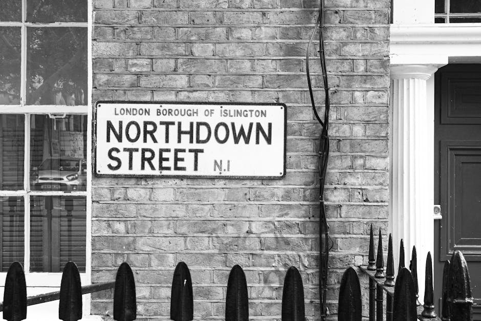 Black and white image showing a brick exterior wall of a building on Northdown Street in the London Borough of Islington, with a street sign attached to the wall reading 'Northdown Street N1'. To the left, there is a window with a white frame and multiple panes, reflecting a parked car and some foliage. Adjacent to the wall on the right, a dark-colored door with decorative paneling is partially visible, surrounded by a white classical-style column on its right. A black metal fence with pointed finials is in the foreground, with some vertical bars blocking part of the view. Extending from the wall, a black cable runs vertically downward, secured with clips. The scene suggests an urban residential setting, possibly during a home relocation process, consistent with the surroundings of house removals and furniture transport services provided by Islington Man and Van.