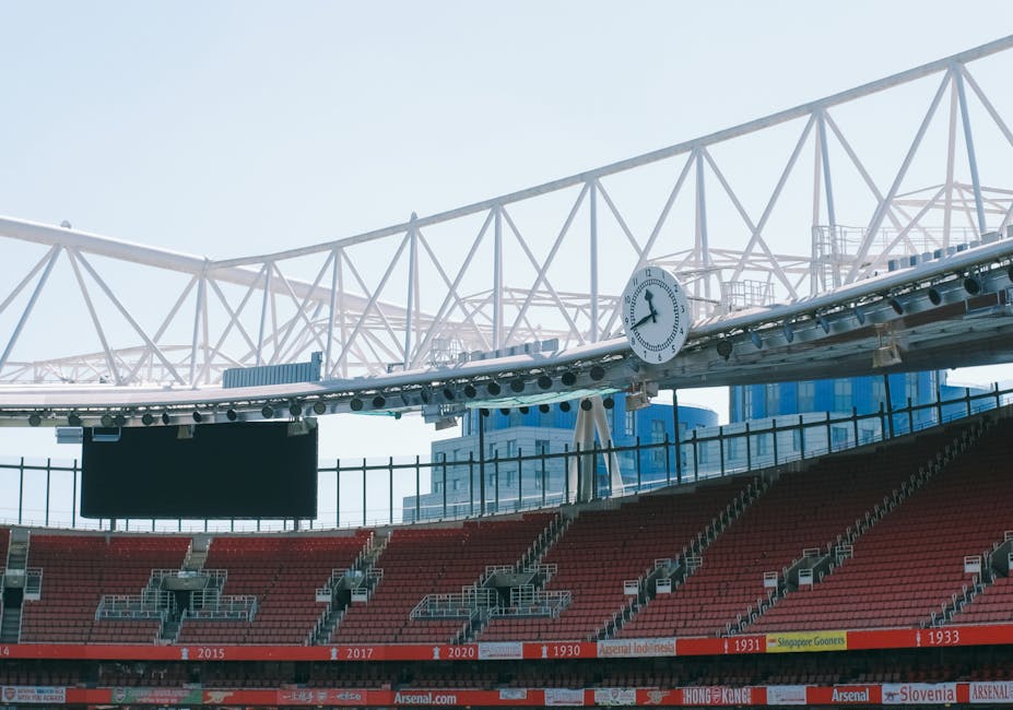 The image shows the interior of a sports stadium with red seating arranged in multiple tiers. A large, black scoreboard is mounted on a railing in front of the seats, and a white clock with black hands and numbers is attached to the stadium's overhead structure, displaying the time as approximately 4:10. The overhead steel framework, painted white, forms a geometric pattern supporting the roof. The stadium is well-lit with natural daylight, and a distant cityscape with tall modern buildings can be seen through the open sides. The seating area appears prepared for spectators, with no fans or players visible. This setting is related to event hosting and sporting logistics, which may involve the delivery or transportation of equipment or furniture for stadium events, similar to home relocation or transportation services offered by Islington Man and Van.