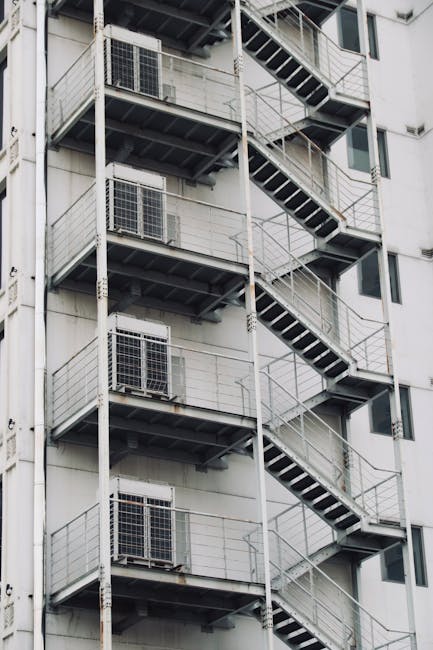 A metal access door with a rusty brown finish is set into a weathered concrete wall on a building's exterior, next to a metal staircase with a teal railing leading upward. The staircase has concrete s