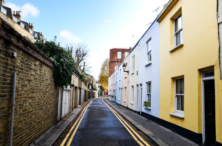 A narrow residential street in Islington with a slight curve, featuring a mix of old brick and modern painted terraced houses on the right side, including buildings painted in pastel yellow, white, and blue. The street surface is wet, and yellow double lines run along the curb. On the left side, a tall brick wall with some greenery and small doors or garages is visible. In the background, there are a few leafless trees and a red brick building, with the sky partly cloudy. This scene illustrates a typical urban setting suitable for home removals involving narrow access, where furniture transport and packing may require careful handling, as managed by local removals specialists like Islington Man and Van.