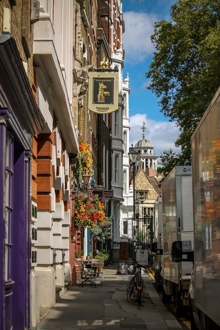 A narrow street scene in Islington showing a row of multi-storey brick and stucco buildings with decorative facades, flower baskets hanging from window sills, and a sign for a pub called 'Young's'. Several cardboard boxes, wrapped in protective plastic and packing materials, are placed on the sidewalk near the entrance of a residential property, indicating packing or home packing preparations. A moving trolleys and a large, white professional removal van are positioned on the pavement, with the van partially loaded or ready for loadings as part of a furniture transport or house removal process. In the background, there is a historic church with a dome and clock tower, under a partly cloudy sky, highlighting the urban context of a house relocation in central London. The setting suggests a step in the loading process for furniture and belongings during a professional removals operation carried out by Islington Man and Van, with visible elements emphasizing narrow access logistics and home moving activities.
