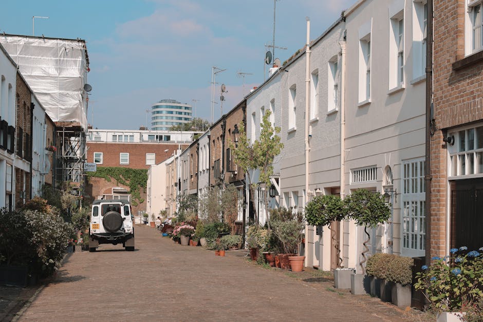 A narrow residential street in Islington with a slight curve, featuring a mix of old brick and modern painted terraced houses on the right side, including buildings painted in pastel yellow, white, and blue. The street surface is wet, and yellow double lines run along the curb. On the left side, a tall brick wall with some greenery and small doors or garages is visible. In the background, there are a few leafless trees and a red brick building, with the sky partly cloudy. This scene illustrates a typical urban setting suitable for home removals involving narrow access, where furniture transport and packing may require careful handling, as managed by local removals specialists like Islington Man and Van.