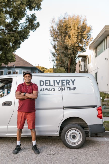 A young man dressed in a red polo shirt, red shorts, and a black cap stands with his arms crossed next to a white delivery van parked on a residential street. The van displays the words 'DELIVERY ALWAYS ON TIME' on its side. Behind him, there are trees with autumn foliage and modern white buildings with large windows, indicating a suburban neighbourhood. The scene is set during daylight with clear weather and natural lighting. This image relates to home relocation and the logistics of furniture transport, which are part of the services offered by Islington Man and Van as they handle packing, loading, and delivery during house removals, including potential delays that can occur in the process.