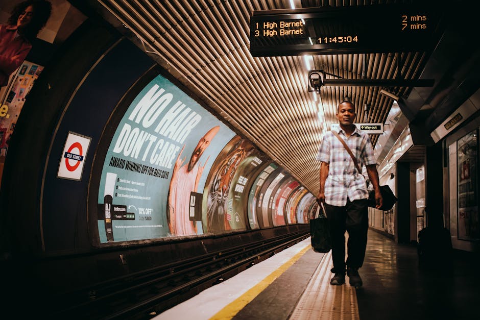 A man carrying a black duffel bag and a small black suitcase is walking along an underground train station platform, likely at the start or end of a home relocation or furniture transport process. The individual is dressed in a light plaid shirt, dark trousers, and appears to be heading towards an exit or a waiting vehicle. Behind him, there are advertisements on the station wall, including a large blue poster with white text, and illuminated digital display boards showing train destinations and departure times, such as High Barnet, with departure times of two and seven minutes. The station features a curved, tiled ceiling with parallel wooden or metal slats, and the platform is equipped with safety markings along the edge. Lighting is bright, highlighting the station's clean, modern environment, suitable for professional removals or moving services provided by Islington Man and Van, who manage various aspects of packing, loading, and transportation in urban settings.
